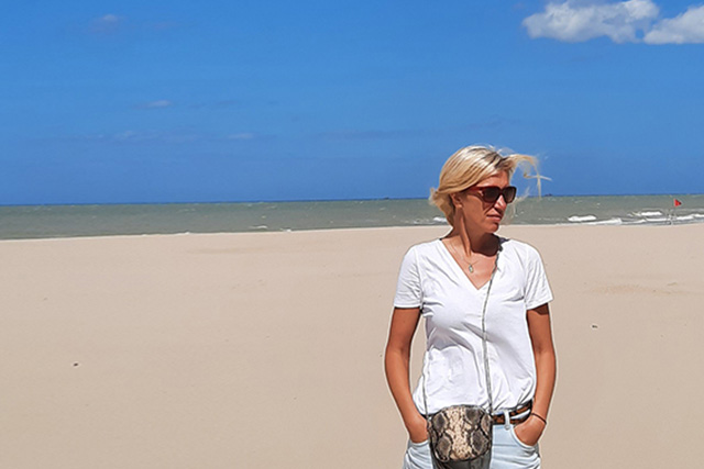 Zdjęcie kobiety w białej bluzce na plaży. W tle morze i błękitne niebo