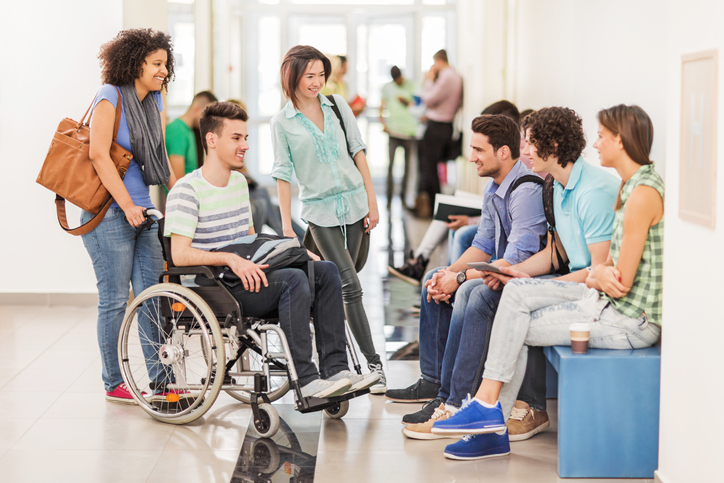 Grupa studentów rozmawiających na korytarzu uczelni. Jeden ze studentów porusza się na wózku inwalidzkim.