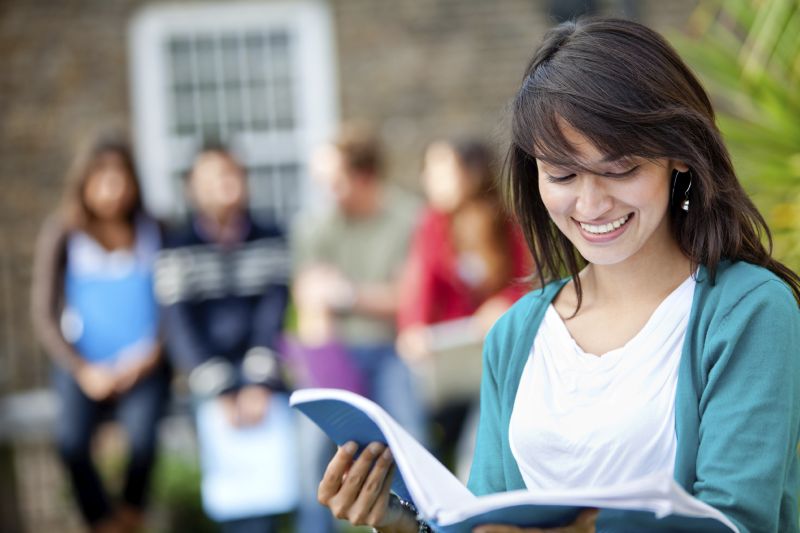 Młoda uśmiechnięta kobieta czyta informacje zawarte w notesie. W tle znajdują się cztery inne osoby.
