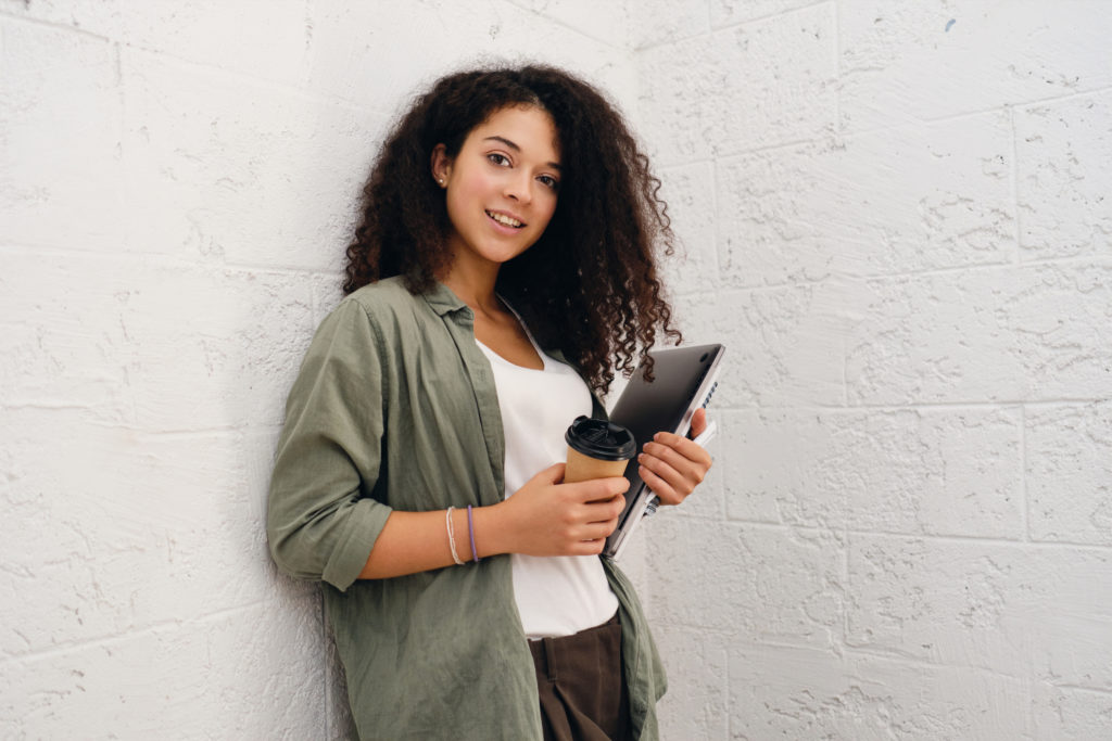 Młoda kobieta z kręconymi włosami opierająca się o ścianę, trzymająca laptop oraz kubek z kawą na wynos