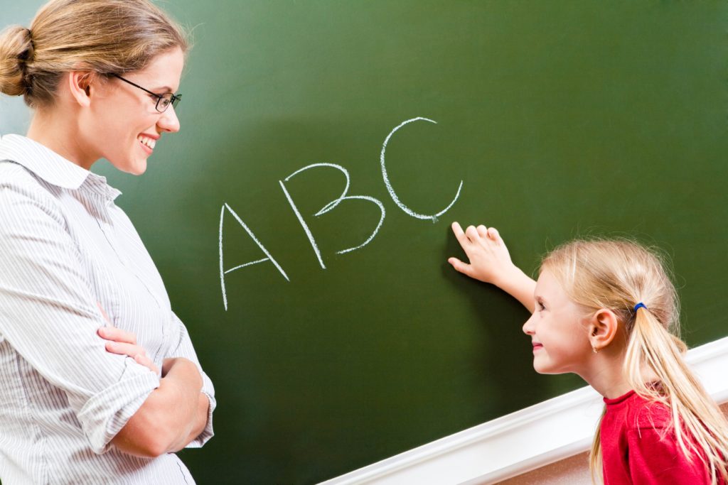 Mała dziewczynka wskazująca nauczycielce napis "ABC" zapisany kredą na zielonej tablicy - obraz symbolizujący kierunek Pedagogika
