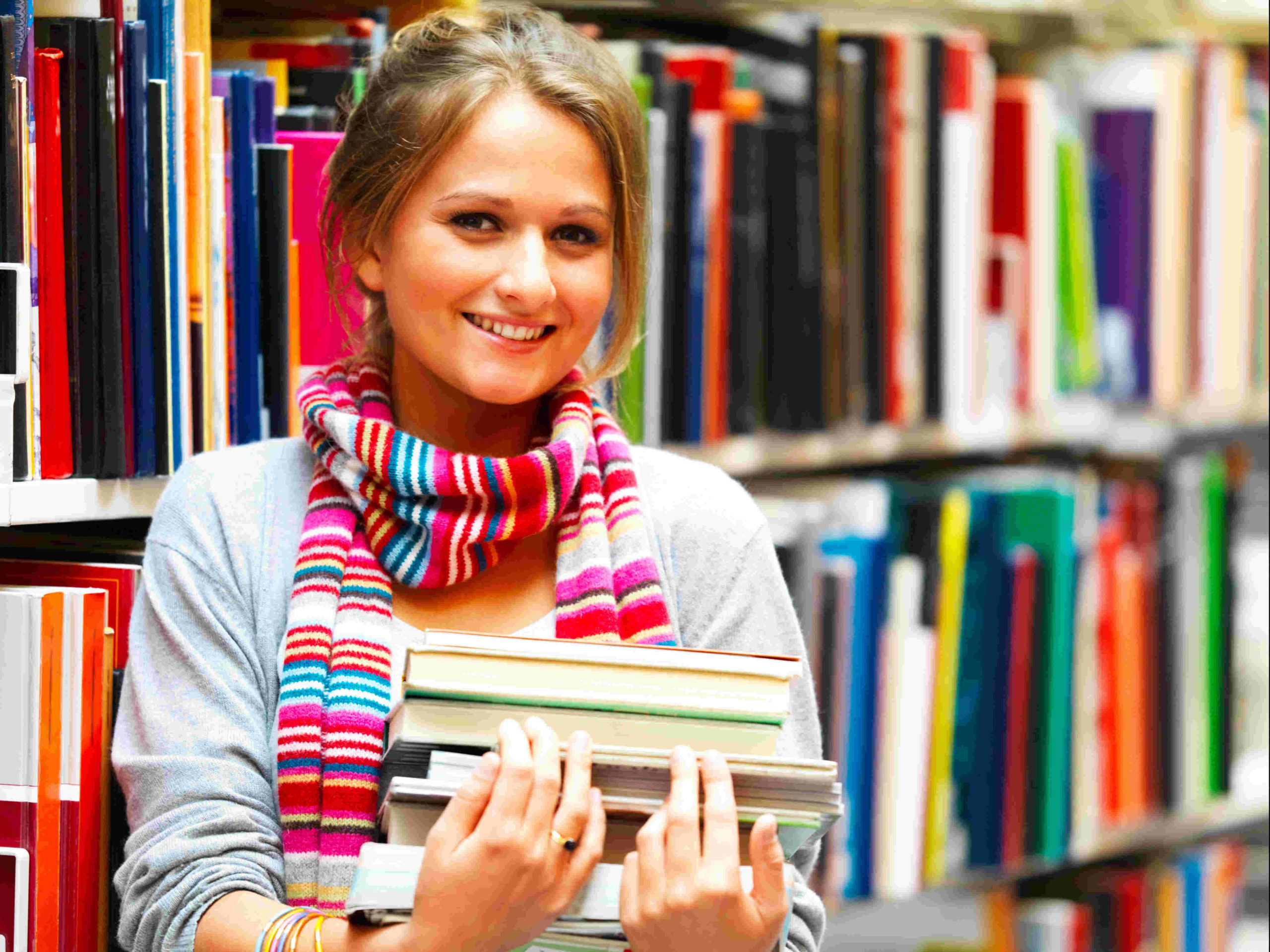 Małoda kobieta ze stosem książek w rękach w bibliotece.
