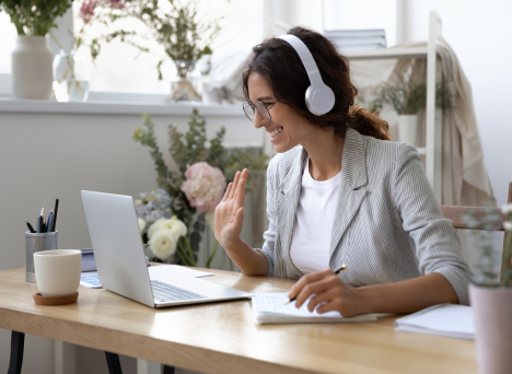 Kobieta siedząca przy biurku na wideokonferennji. Ma uniesioną prawą dłoń, w lewej trzyma długopis, którym zapisuje notatki. Na uszach założone ma białe słuchawki.