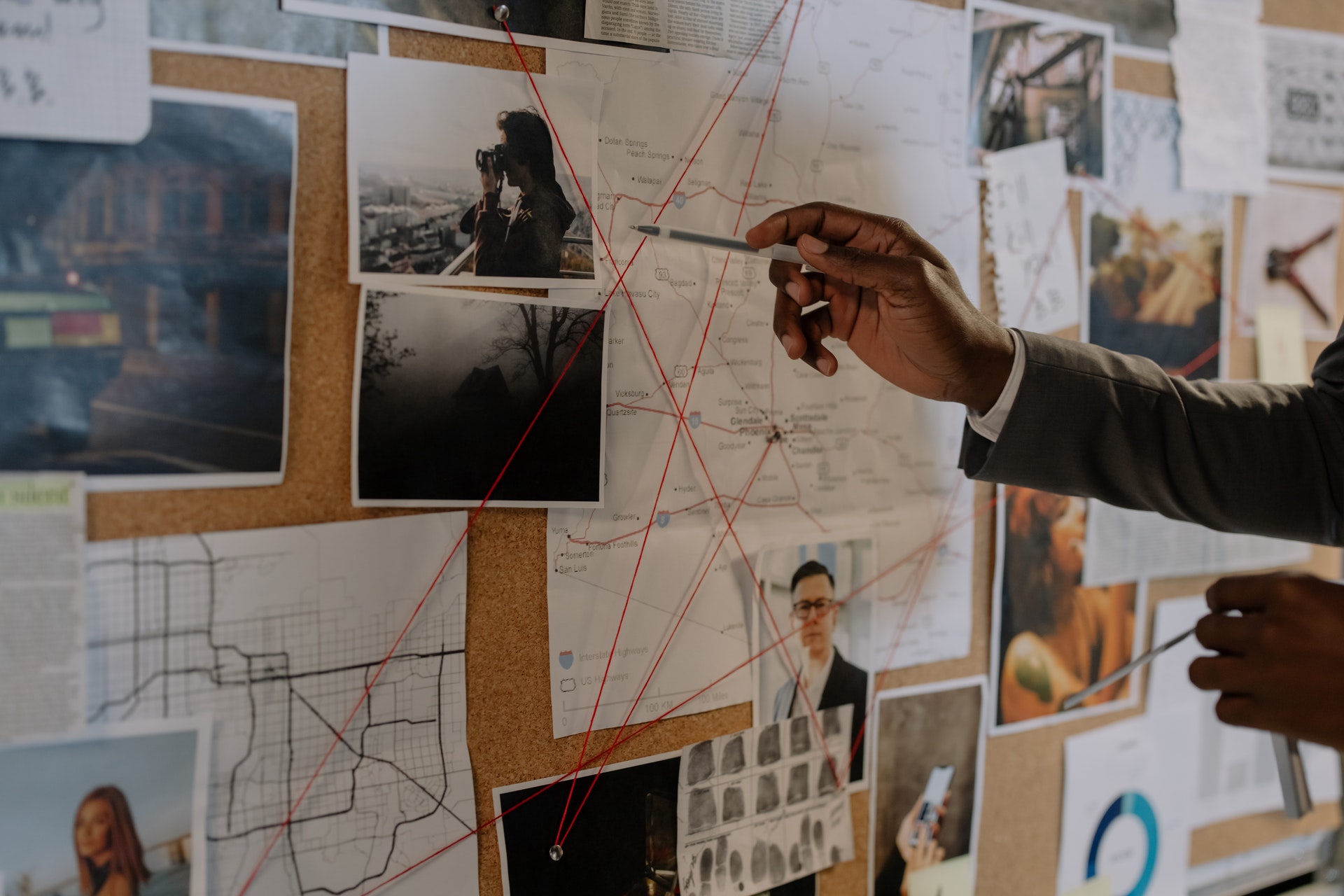 Tablica korkowa ze zdjęciami oraz mapami, połączonymi ze sobą czerwoną linką. Dłoń mężczyzny przy użyciu długopisu wskazuje miejsca na mapie.