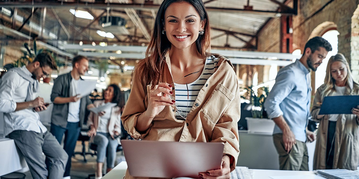 Uśmiechnięta kobieta siedząca przy biurku z laptopem, w prawej dłoni trzyma okulary. W tle znajdują się inne osoby pracujące w tym samym miejscu.