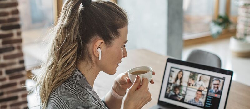 Kobieta z kubkiem kawy i słuchawkami w uszach na spotkaniu online. W tle znajduje się laptop z widoczną konferencją na ekranie.