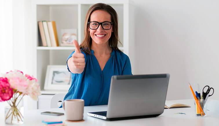 Młoda kobieta w okularach siedząca przy biurku z laptopem. Jej prawa dłoń jest podniesiona do góry i wskazuje symbol 