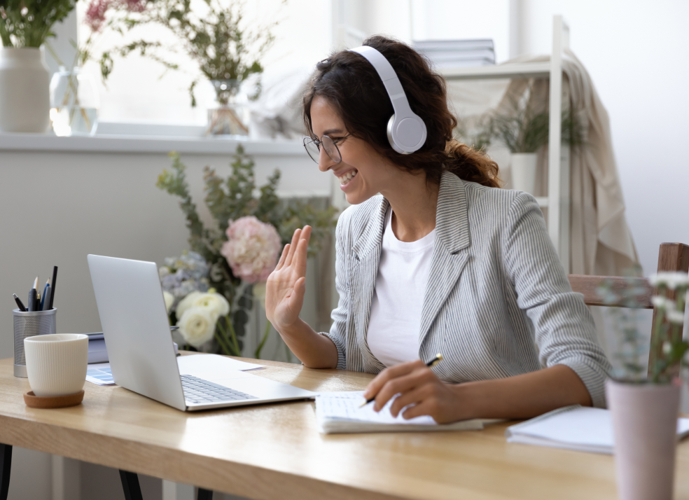 Kobieta siedząca przy biurku na wideokonferennji. Ma uniesioną prawą dłoń, w lewej trzyma długopis, którym zapisuje notatki. Na uszach założone ma białe słuchawki. 