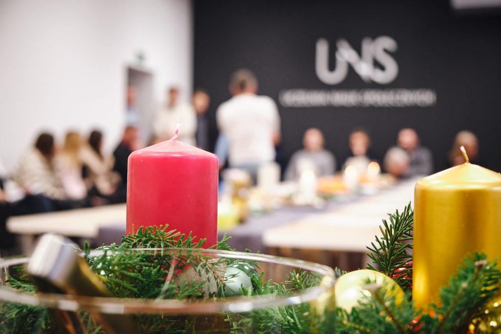 Zdjęcie świecy i dekoracji bożonarodzeniowych, w tle grupa studentów siedząca pod ścianą z logotypem Uczelni Nauk Społecznych