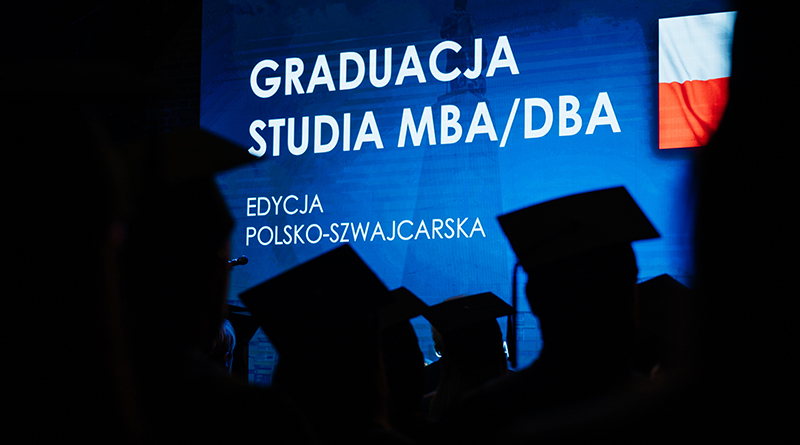 Zdjęcie osób w akademickich biretach na tle tablicy LED z grafiką